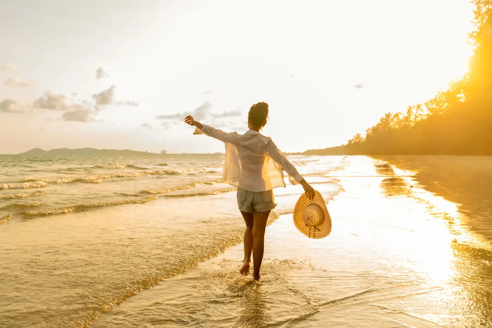 Backlit walker holding straw hat on beach